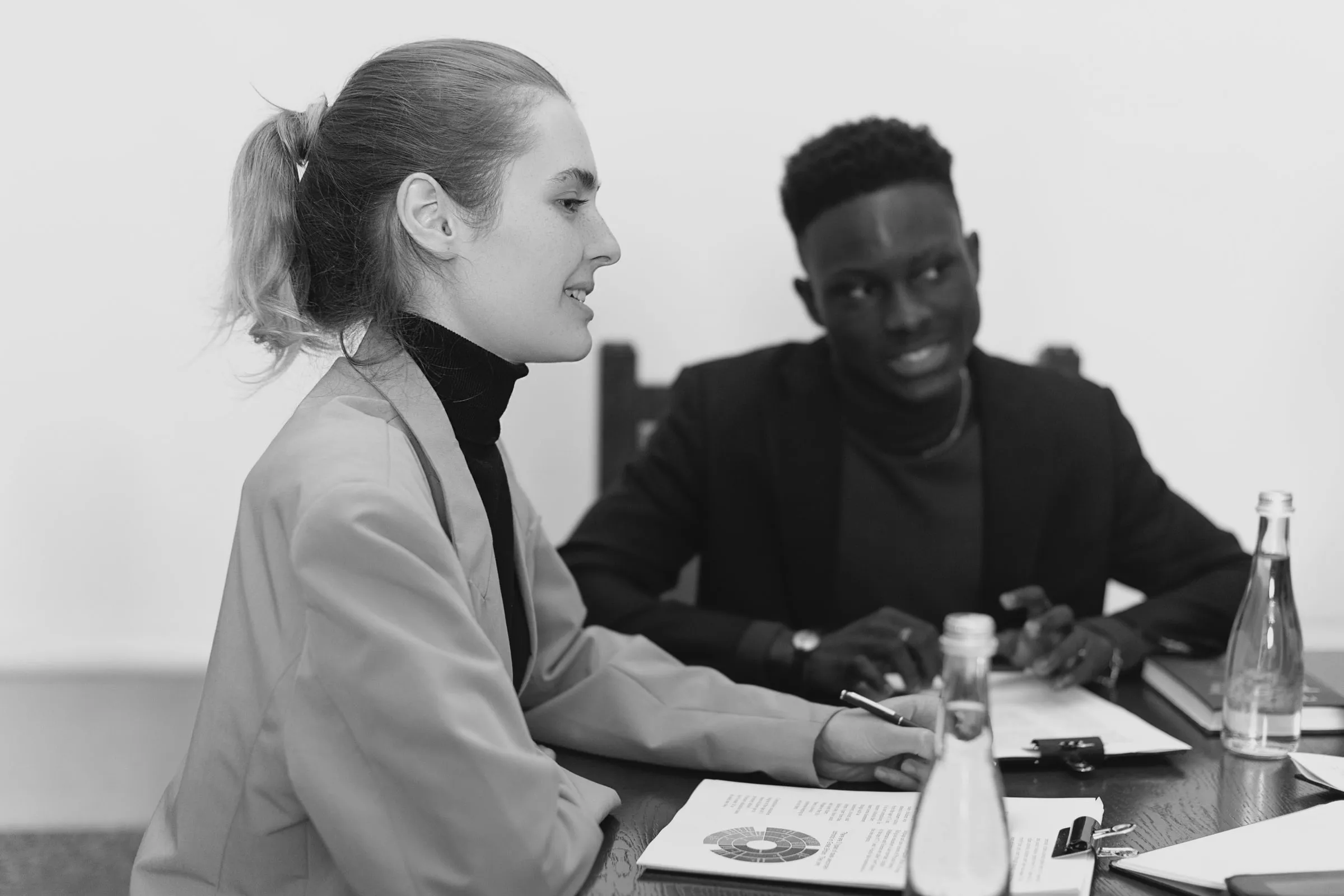 A conversation at a meeting table: a woman with her hair tied back speaks, a pen in her hand over papers, while a colleague in a dark jacket listens across the table with a smile.