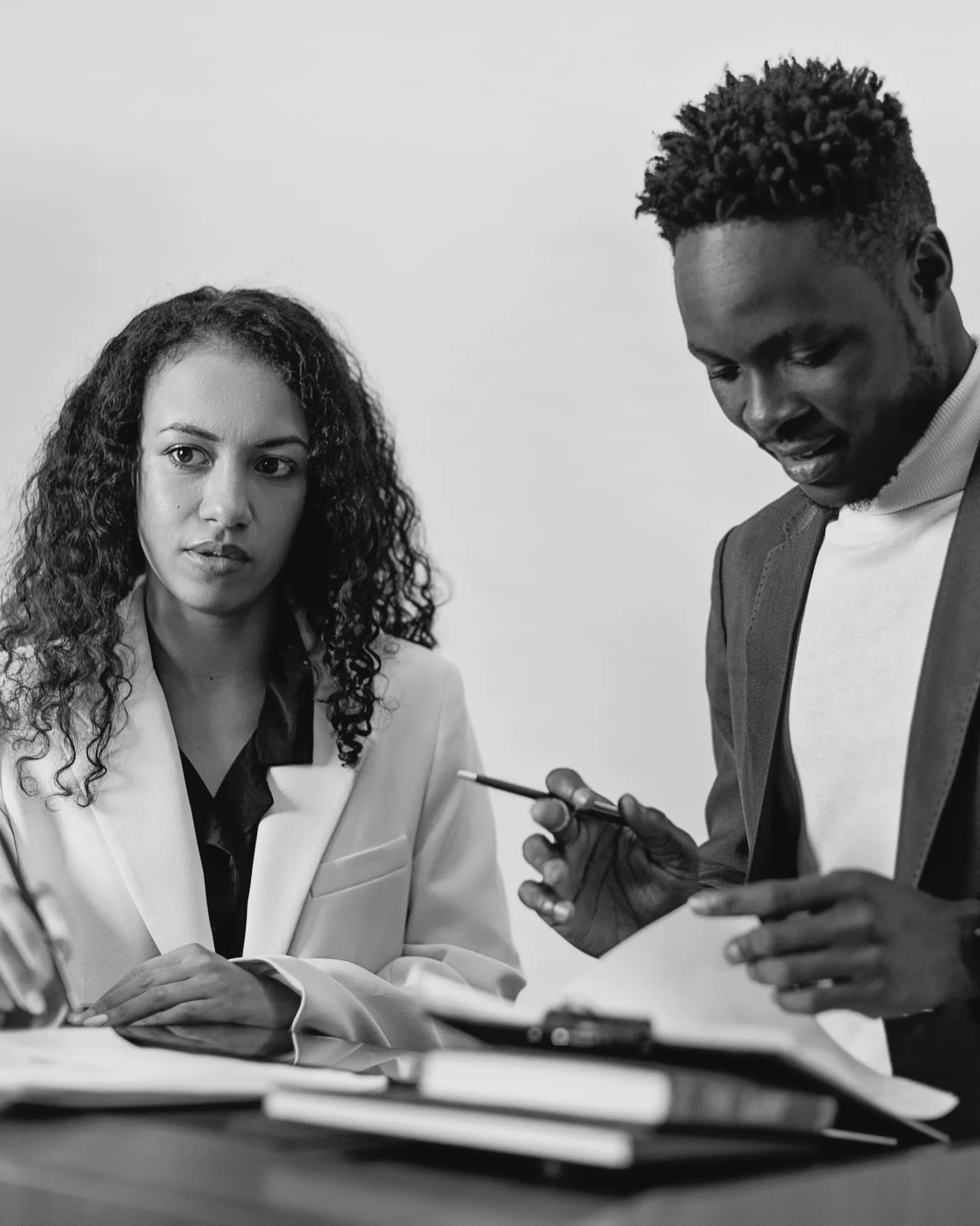 Two lawyers at a desk: a woman with long curly hair, in a light blazer, looks up from her papers with a pen in hand, while her colleague beside her, in a grey blazer and white turtleneck, reads a page with a pen in his hand.